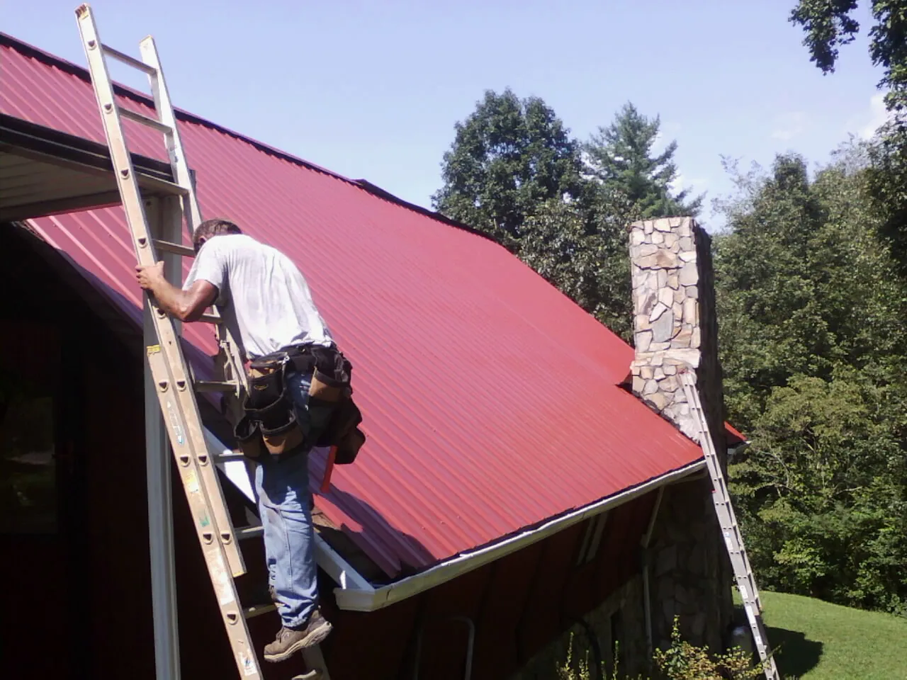 Roofer on a ladder beside a red metal roof and stone chimney — Soffit & Fascia Repair by Roofing Brothers in Outlook